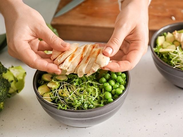 Händer lägger skivat kycklingkött på en skål med gröna grönsaker